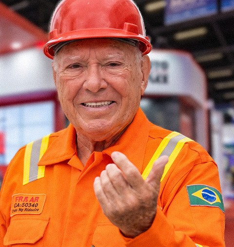Célio Couto, de macacão laranja e capacete vermelho, sorrindo e fazendo sinal para você se aproximar do estande da CM Couto.